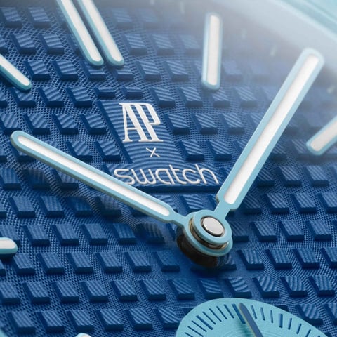 Close-up of a Swatch watch face showing AP and Swatch branding with silver hour markers on a blue textured dial